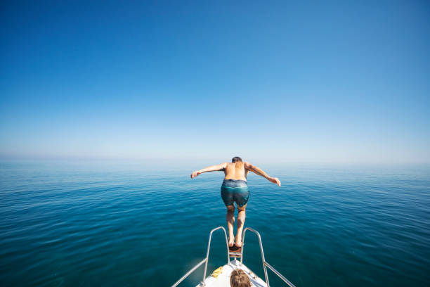 Muscular mid adult man diving into the sea water from deck of anchored yacht or sailboat. He is wearing red swimwear which makes gread contrast with surrounding colors. Photo taken from sea surface level.