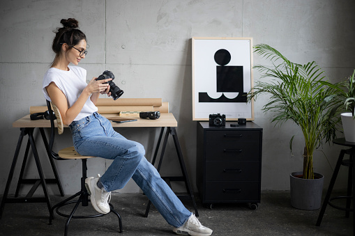 Side view of a young confident Caucasian female photographer sitting on a chair at home, relaxing and taking a break from work while tinkering with her photographic camera equipment