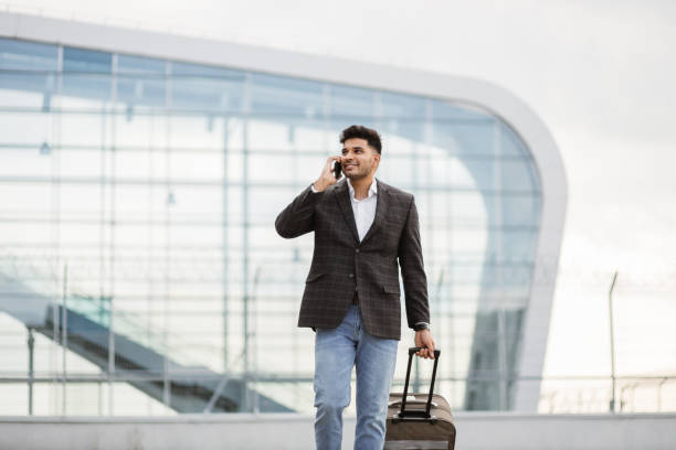 uomo d'affari indiano che cammina fuori alla stazione o all'aeroporto parlando al telefono e portando la valigia - travel people traveling business travel vacations foto e immagini stock