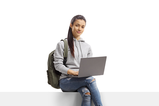 Female student with a backpack sitting on a blank panel with a laptop isolated on white background