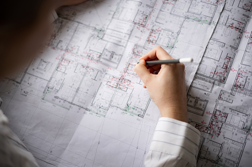 Close up view of an unrecognizable professional young Caucasian female architect analysing and drawing on a blueprint in front of her in her office, concentrating on her work