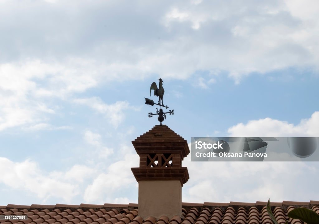 Weather vane in the form of a rooster on the roof. Weather vane in the form of a rooster on the roof. Shows where is East, where is West,"nSouth and North. Thanks to this device, the weather vane will determine not only the"nstrength of the wind, but also a fairly accurate direction in one direction or another."r Rooster Stock Photo