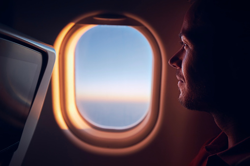 Portrait of man traveling by airplane. Passenger looking through plane window during flight at sunrise.