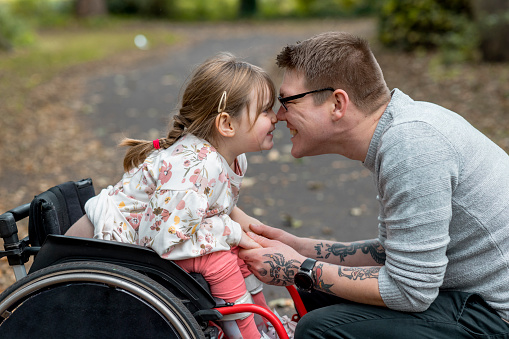 A close up side view of a father and his young daughter who is a wheelchair user having a cute affectionate moment with each other whilst on a family day out in a beautiful public park in Newcastle upon Tyne in the North East of England.