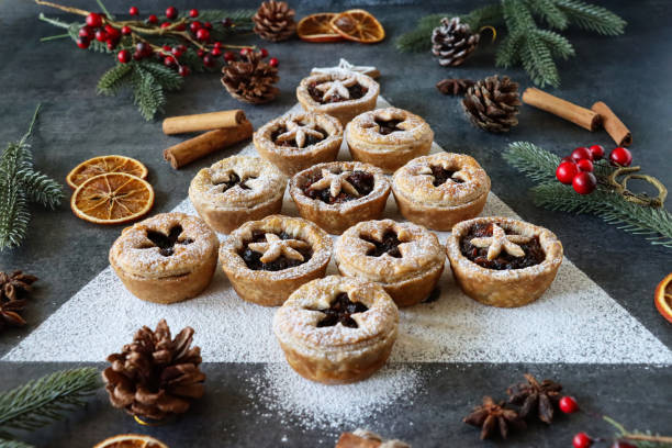 istock Close-up image mince pies laid out like Christmas tree surrounded by decorations, festive Christmas dessert, cinnamon stick bundle, red berries, spruce needles, pine cones, star anise, mottled grey background, focus on foreground 1384244899
