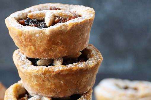 Stock photo showing close-up view of a stack of individual mince pies made with homemade short crust pastry glazed with an egg wash. The pies have been topped with pastry star detail, hiding the sweet mincemeat (mixture of chopped dried fruit, distilled spirits and spices) filling.