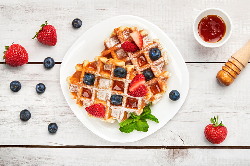 White plate with Belgian waffles with fresh strawberries, blueberries, mint leaves, honey and honey dipper beside. White wooden background, top shot