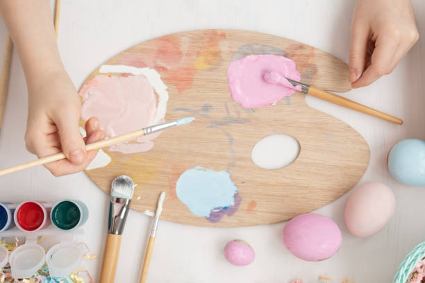istock Preparation for Easter. The child is holding a large egg in his hands. Pastel shades on the palette. A child's hand paints an egg light blue with a brush. Happy Easter 1384249016