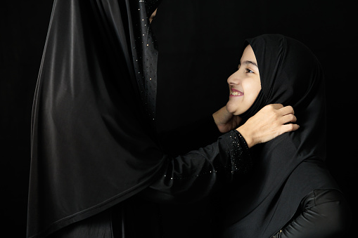 muslim woman and her daughter smiling and hugging together on black background
