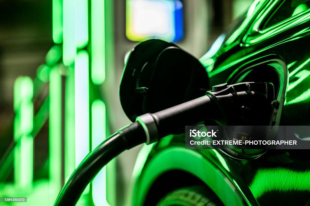 Electric car charger and green reflection from the light Close-up on an electric car charger plugged into car on public parking with electric charging stations. Electric Vehicle Stock Photo