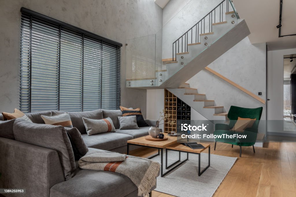 Stylish composition of modern living room design with corner grey sofa, green armchair, coffee table and minimalist personal accessories. Stairs in living room interior. Concrete walls. Template. Decorating Stock Photo