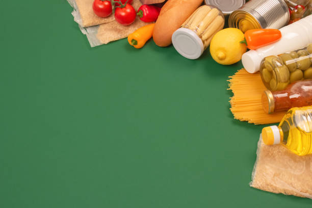 istock Different groceries, food donations on green background with copy space - pasta, vegatables, canned food, baguette, cooking oil, tomatoes. Food bank or food delivery concept, support refugees. Selective focus 1384256828