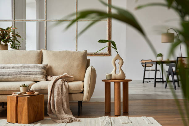 istock Modern living room interior design with beige sofa, wooden side table, plants and creative accessories. Template. Copy space. Dining room in the background. 1384257129