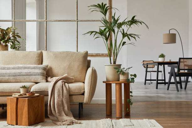 istock Modern living room interior design with beige sofa, wooden side table, plants and creative accessories. Template. Copy space. Dining room in the background. 1384257735