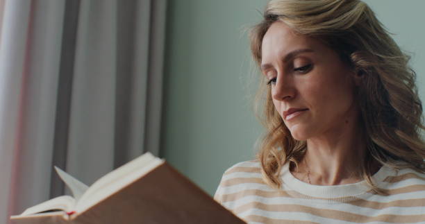 istock Female student reads a book in a cozy bright living room, sitting on a sofa. Learning, studying, education, read books, stay home concept. 1384264283