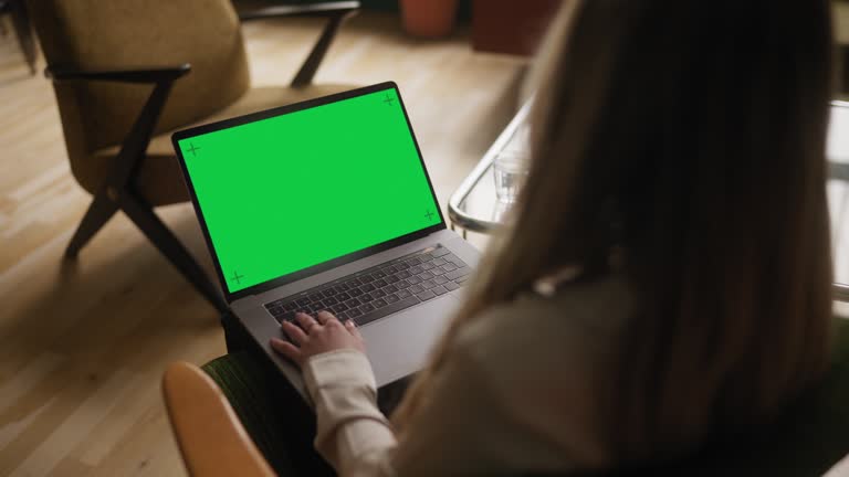 Young woman working from home. She is sitting in a vintage armchair in her apartment, working on her laptop. Chroma key green screen on the laptop.