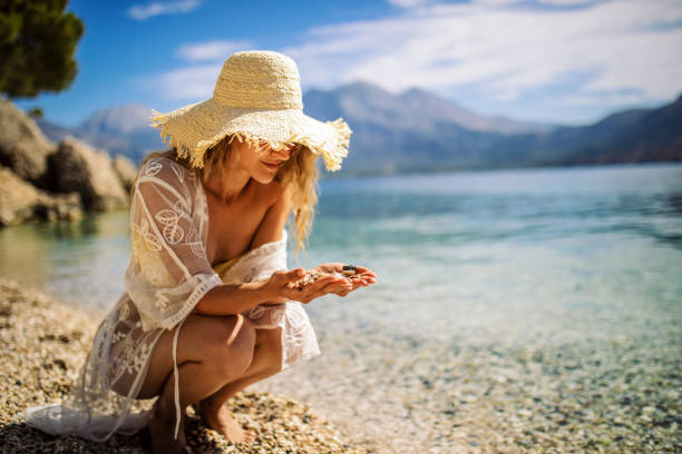 Attractive young woman enjoying day on beach where she collects shells and pebbles. She wears bikini with transparent cover and hat.