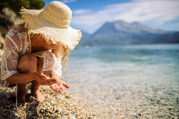 Attractive young woman enjoying day on beach where she collects shells and pebbles. She wears bikini with transparent cover and hat.