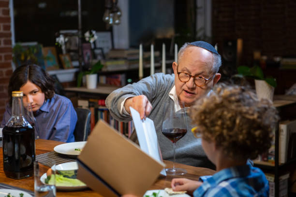 Grandfather Helping Little Boy Read from Haggadah at Pasover Seder A modern Jewish American family celebrates Passover together. The youngest person at the seder, a little boy, is asking the Four Questions (Mah Nishtanah). These questions provide the impetus for telling why this night is different from all other nights. passover celebration stock pictures, royalty-free photos & images