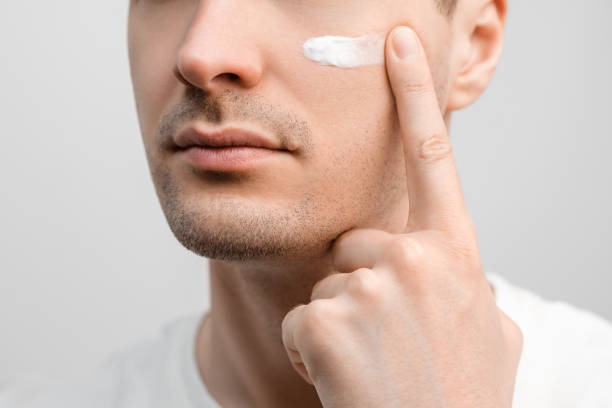 Handsome man with a stubble in a white t-shirt, sample of a face cream on his cheek, smudging with finger. stock photo