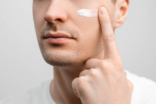 handsome man with a stubble in a white t-shirt, sample of a face cream on his cheek, smudging with finger. - applied science imagens e fotografias de stock