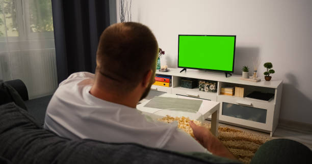 istock Man Watching Television with Green Screen Chroma Key, shot behind models shoulders. Man Eating Popcorn Relaxing Sitting on a Couch Home looking to TV. 1384325634