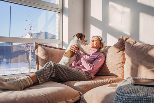 Adult pretty woman in casual clothes hugging her  dog sitting on the sofa in living room of her cozy country house. Animal communication concept