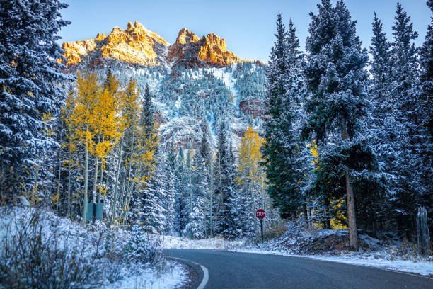 Photo of Maroon Bells Creek road in Aspen, Colorado rocky mountains covered in snow after winter late fall snowfall with sunlight on peak and dark forest yellow trees