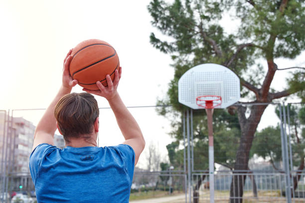 istock Young player throwing to the basket. Unrecognizable Caucasian man with his back turned playing on a basketball court. 1384344496