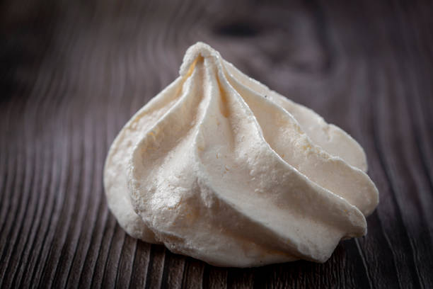 Flat lay of homemade meringue cookies arranged on a cooling rack with natural baking ingredients, including eggs, cinnamon sticks, star anise, dark chocolate pieces, and granulated sugar on a rustic blue wooden background. The scene captures a slow, sustainable baking process with simple, real ingredients and warm, cozy textures. Copy space appears along the lower portion of the image.