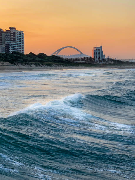 Beautiful warm sunset view over the Moses Mabhida Sadium from the water in Durban, South Africa. View from the blue waters of the Indian Ocean to the deep orange sunset sky with the famous Moses Mabhida Sadium arch in distant background. zululand stock pictures, royalty-free photos & images