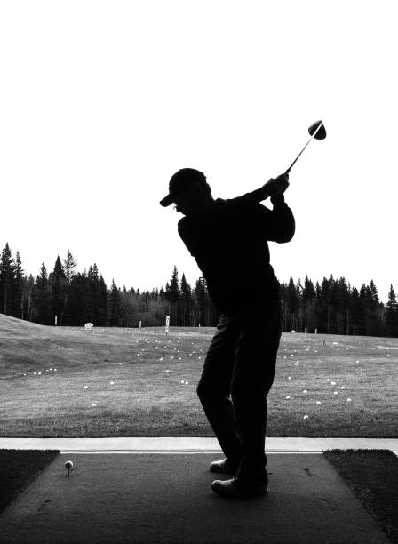 istock Black and white silhouette of man practicing golf at a driving range standing in practice facility with golf range, range balls and forest in the background. 1384350691