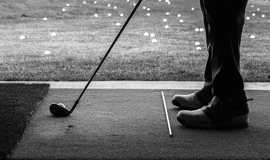Black and white silhouette of mans feet and practice turf with golf club and practice rod as he practices golf at a driving range. Taken from inside the practice facility looking out to the range littered with practice golf balls.