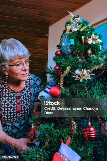 A Grey Haired Senior Latin American Lady Is Putting In The Finishing Touches To To Decorating An Artificial Christmas Tree In Her Apartments Living Room With Great Concentration For The Family Stock Photo - Download Image Now