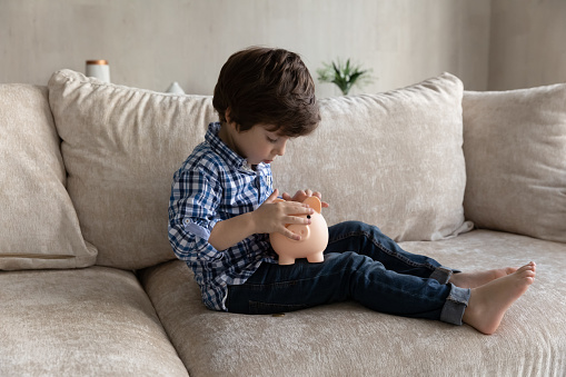 Preschool 6s cute boy sitting on sofa alone in living room holding piggy bank counting and throwing coins into thrift-box. Learns frugality, save funds, keep pocket money for future purchase concept