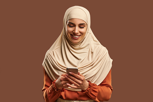 Beautiful Muslim woman in a beige hijab holds a phone in her hands and smiles. Advertising, technology, devices.
