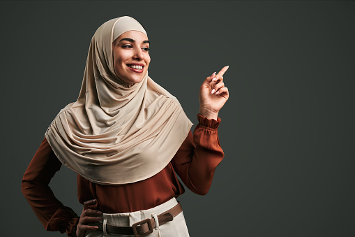 Beautiful muslim woman in beige hijab pointing finger at space on gray background. Copy space.