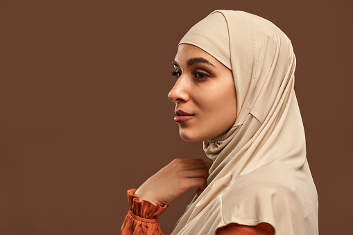 Close-up portrait of beautiful muslim woman in beige hijab on brown background. The woman looks away.Copy space.