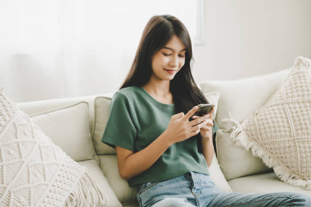 счастливая молодая азиатская женщина, отдыхая дома, она сидит на диване и использует мобильный смартфон - smart phone asian ethnicity mobile phone women стоковые фото и изображения