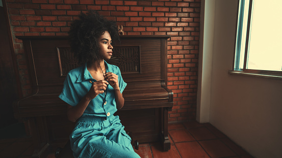 View of a ravishing young Brazilian female with curly afro hair sitting on the chair in the room and pensively looking outside the window with a piano behind her and a copy space place on the right