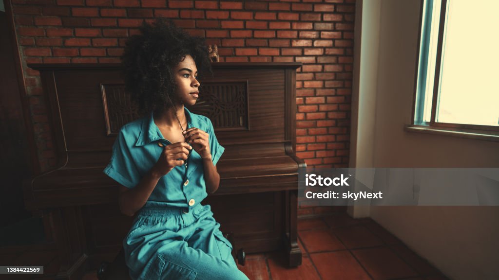 Thoughtful black girl next to piano View of a ravishing young Brazilian female with curly afro hair sitting on the chair in the room and pensively looking outside the window with a piano behind her and a copy space place on the right People Stock Photo