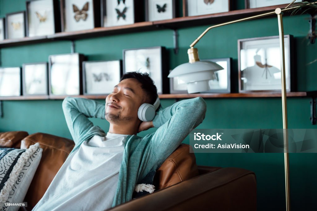 Young Asian man with hands behind head, relaxing on sofa and listening to music with headphones at home. Relaxed young man lying on sofa with music. Relaxing lifestyle, people and technology concept Relaxation Stock Photo