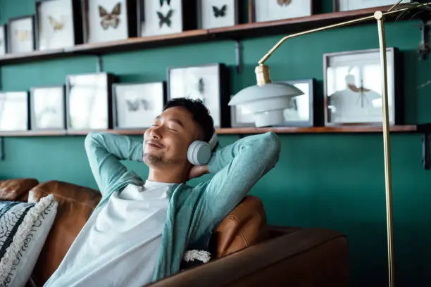Photo of Young Asian man with hands behind head, relaxing on sofa and listening to music with headphones at home. Relaxed young man lying on sofa with music. Relaxing lifestyle, people and technology concept