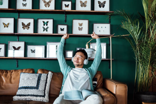 Young Asian man listening to music with headphones, stretching arms while reading book in living room at home. Taking a break. Relaxed young man resting on sofa. Relaxing lifestyle, people and technology concept joven asiático escuchando música con auriculares, estirando los brazos mientras leía un libro en la sala de estar de su casa. tomar un descanso. joven relajado descansando en el sofá. estilo de vida relajante, personas y concepto de tecnología