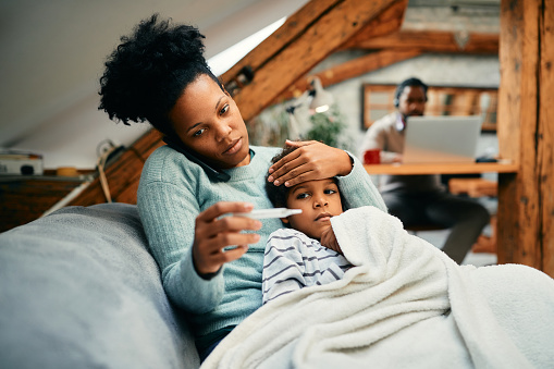 Worried black mother talking on the phone while measuring son's temperature at home.