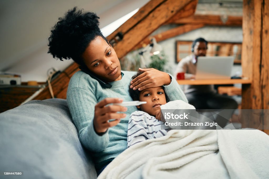 Worried black mother talking on the phone while measuring son's temperature at home. African American mother measuring sick son's temperature while talking to the doctor over the phone. Cold And Flu Stock Photo