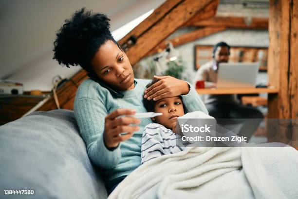 Worried Black Mother Talking On The Phone While Measuring Sons Temperature At Home Stock Photo - Download Image Now