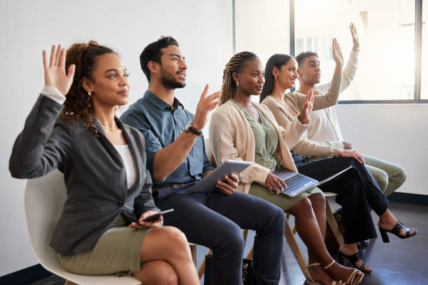 shot of a group of young businesspeople raising their hands while sitting in a row in a modern office - gesturing interview business sitting imagens e fotografias de stock