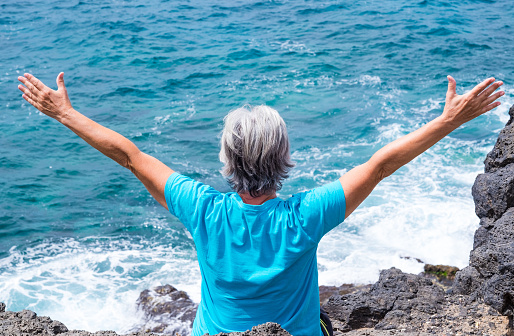 Mature caucasian woman with arms wide open standing on rock and looks to ocean