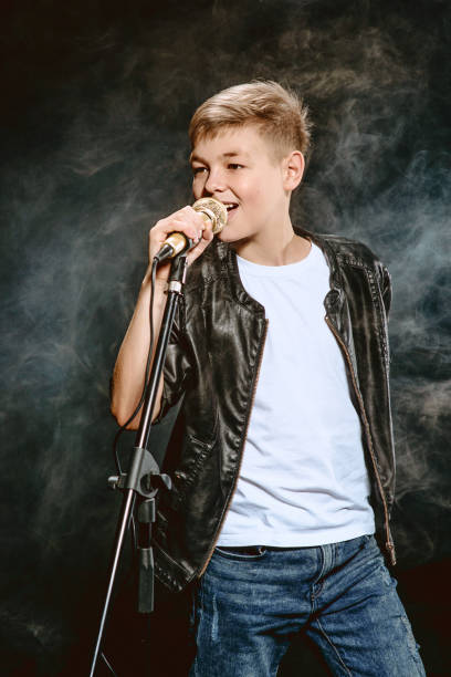 retrato de un adolescente caucásico con camiseta blanca, jeans azules y chaqueta de cuero con micrófono cantando sobre fondo oscuro. concepto de pasatiempo y gloria - singing singer teenager contest fotografías e imágenes de stock